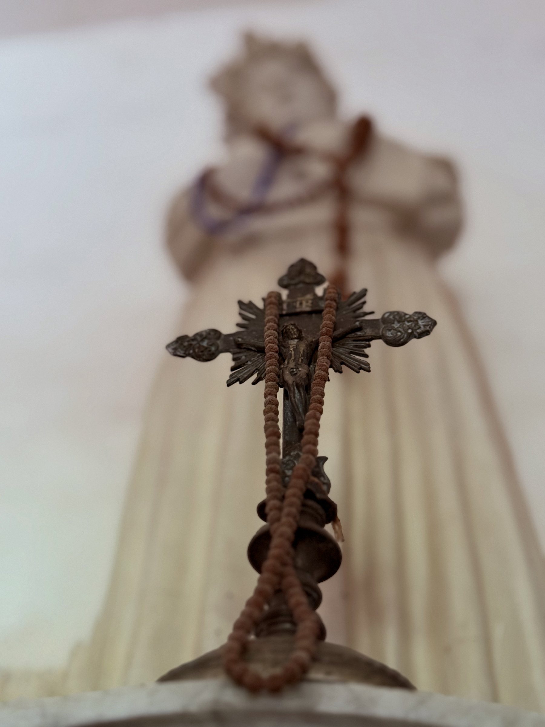 A bronze crucifix with a wooden rosary before a white statue of the Virgin in the Chapelle Saint-Saturnin, Siagne gorge