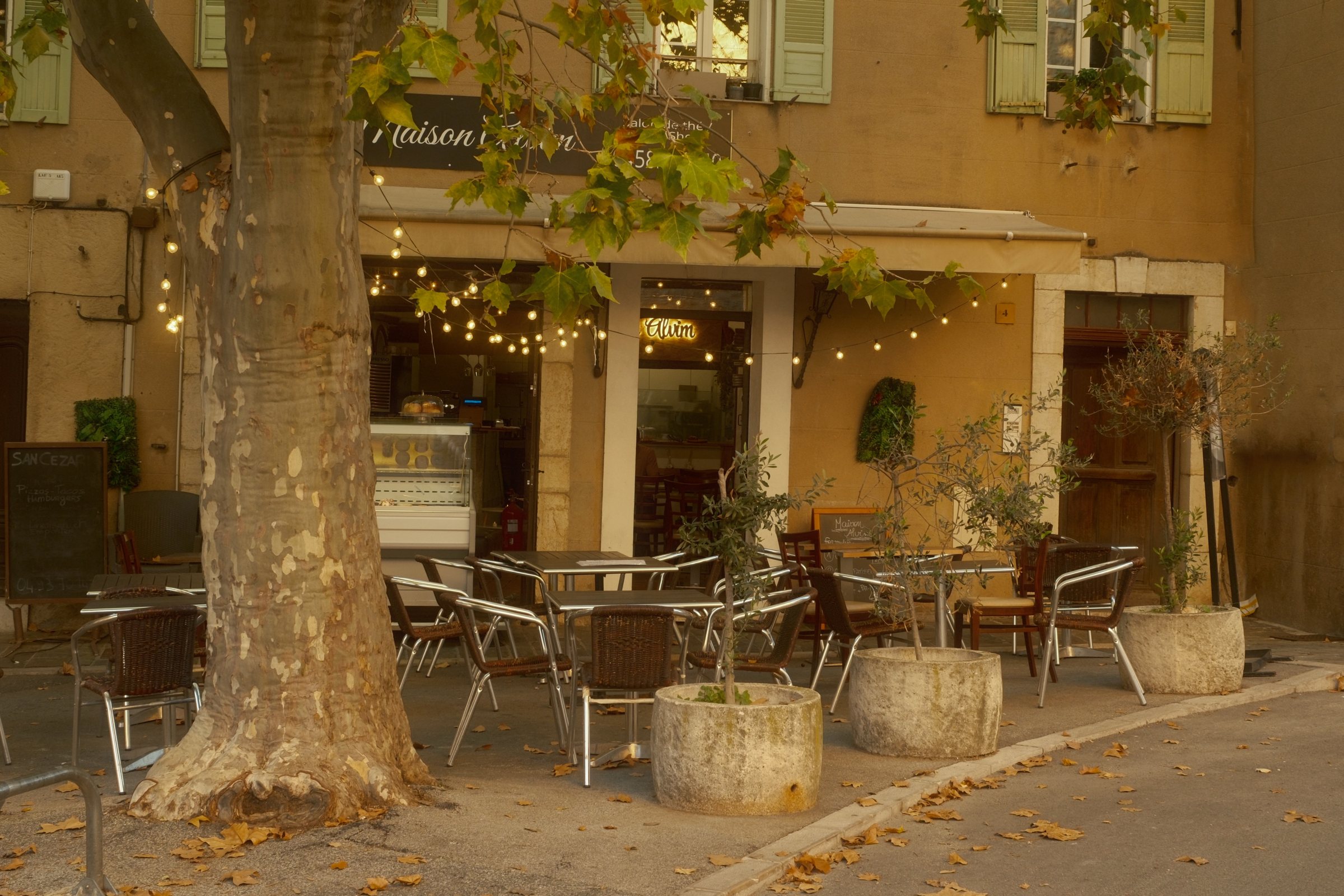 Café terrace under a plane tree on the village square of Saint-Cézaire-sur-Siagne in evening light, chairs empty, fairy lights on